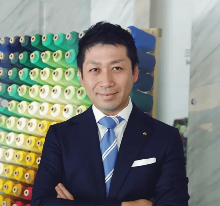 紡錘のカラーバリエーションを背景に、代表取締役社長が腕組している写真